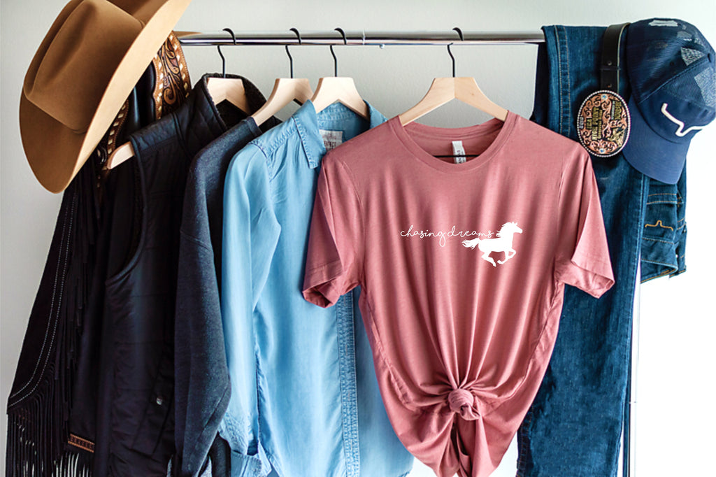 dusty rose pink short sleeve tshirt printed with chasing dreams and a silhouette of a horse in white ink.  Shown with cowgirl hat and jeans hanging on a clothing rack